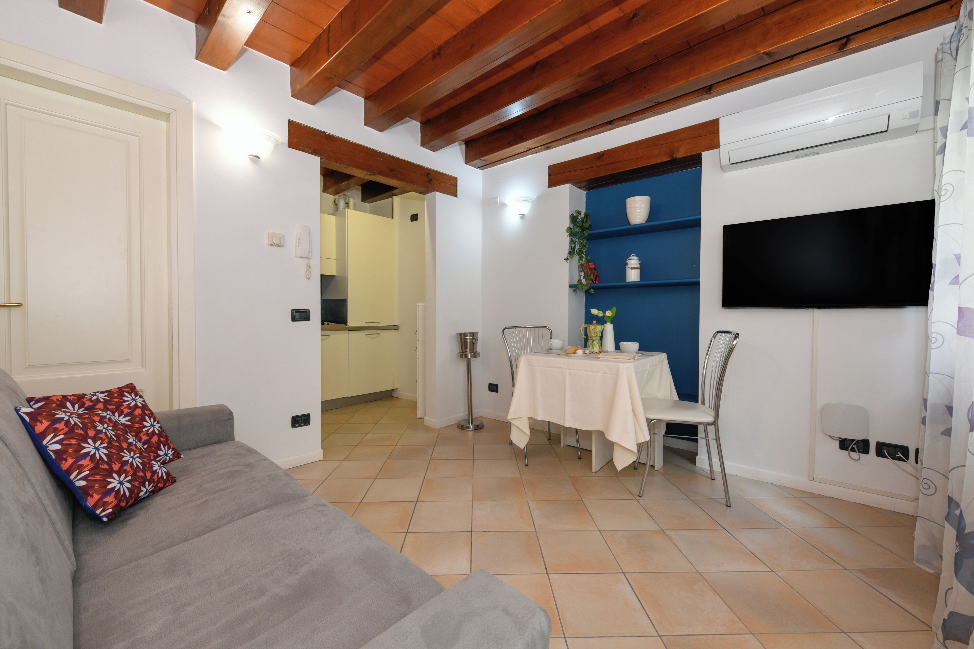 A living room with a sofa, a dining table with chairs, a TV mounted on the wall, and an air conditioning unit.