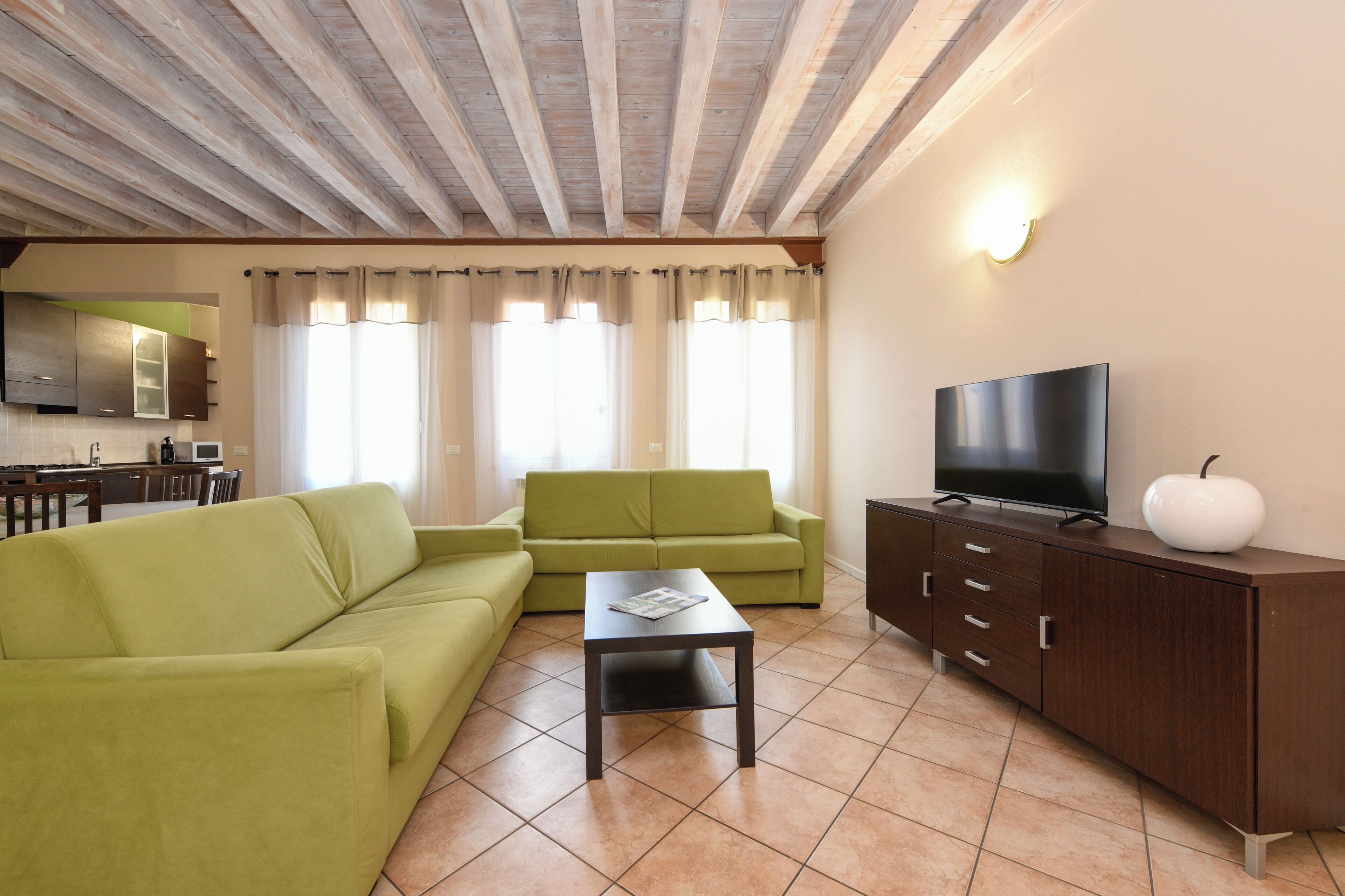 A living room with a green sofa, a coffee table, a TV on a wooden cabinet, and a kitchen area in the background.