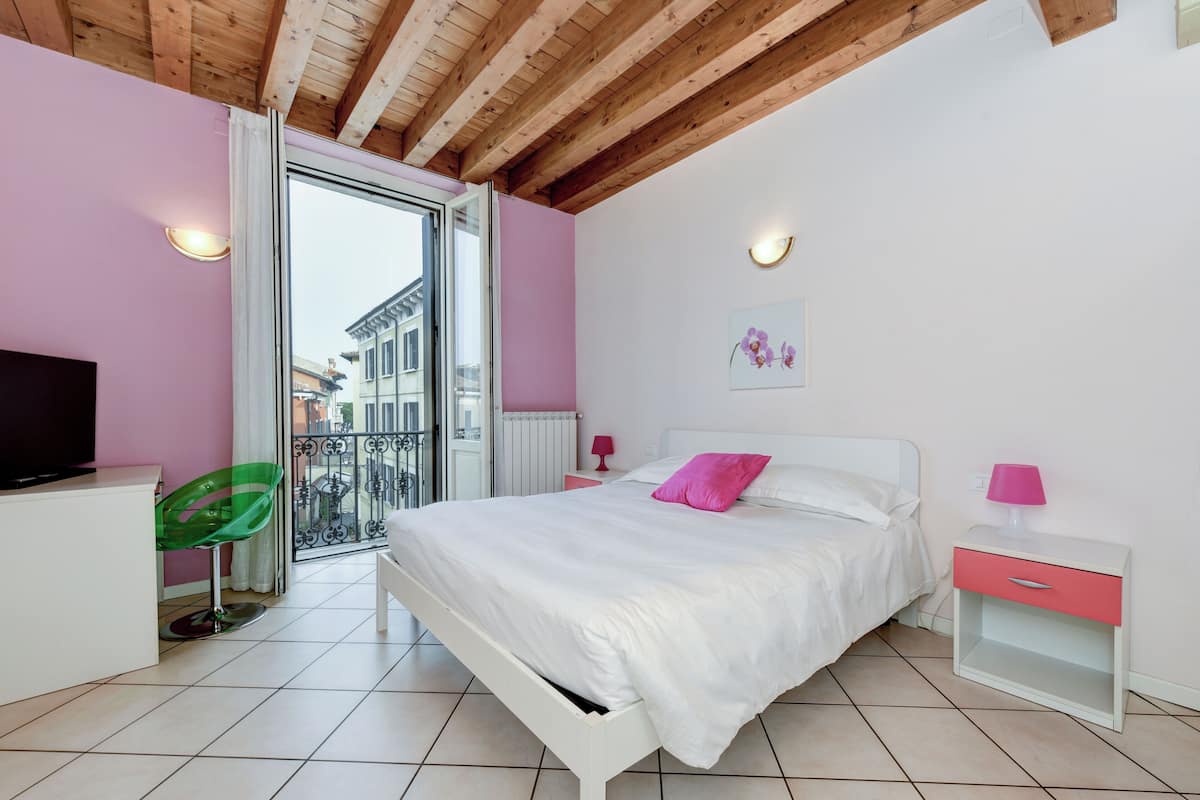 Ein Schlafzimmer mit Bett, Fernseher, Sessel, Nachttisch und einem Fenster mit Blick auf Gebäude.