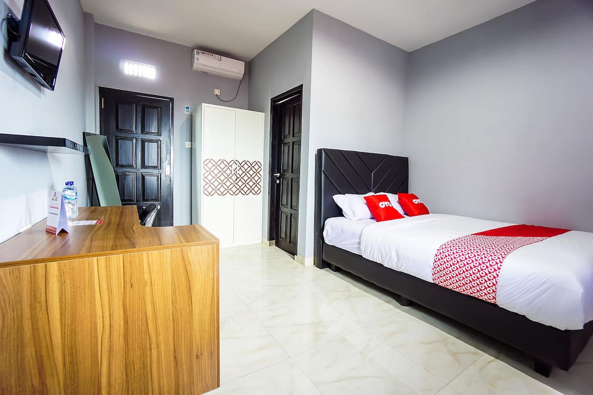 Quarto de hotel com uma mesa de recepção de madeira, uma cama com roupa de cama branca e almofadas vermelhas, uma cabeceira preta e uma parede cinza.