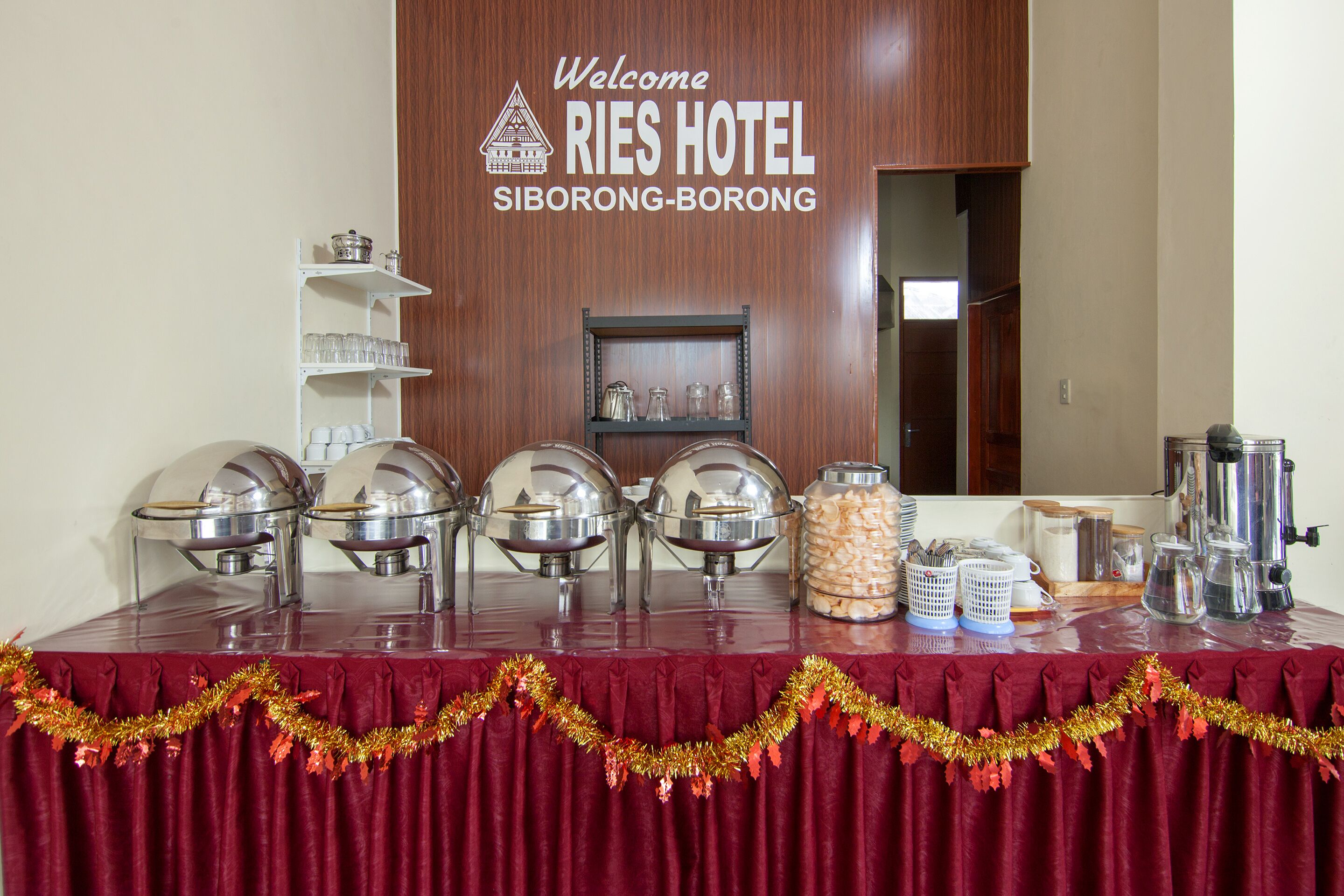 A buffet setup with multiple serving stations, a welcome sign for RIES HOTEL in Siborong-Borong, and a variety of food containers.