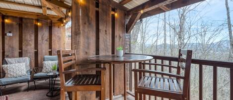 A wooden deck with a table and chairs, a potted plant, and a view of trees and mountains.