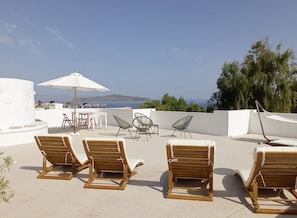 Um terraço no telhado com espreguiçadeiras de madeira, uma mesa e cadeiras sob um guarda-sol branco, com vista panorâmica para o mar e colinas ao longe.