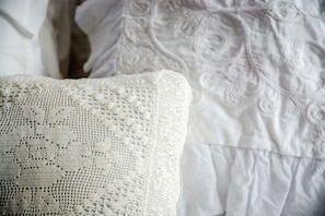 Close-up de um travesseiro branco, com um bordado de crochê detalhado, e um cobreleito branco com um padrão floral discreto.