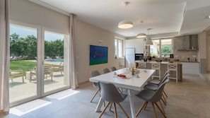 A spacious dining area with a large table and chairs, a view of a swimming pool, and a modern kitchen in the background.