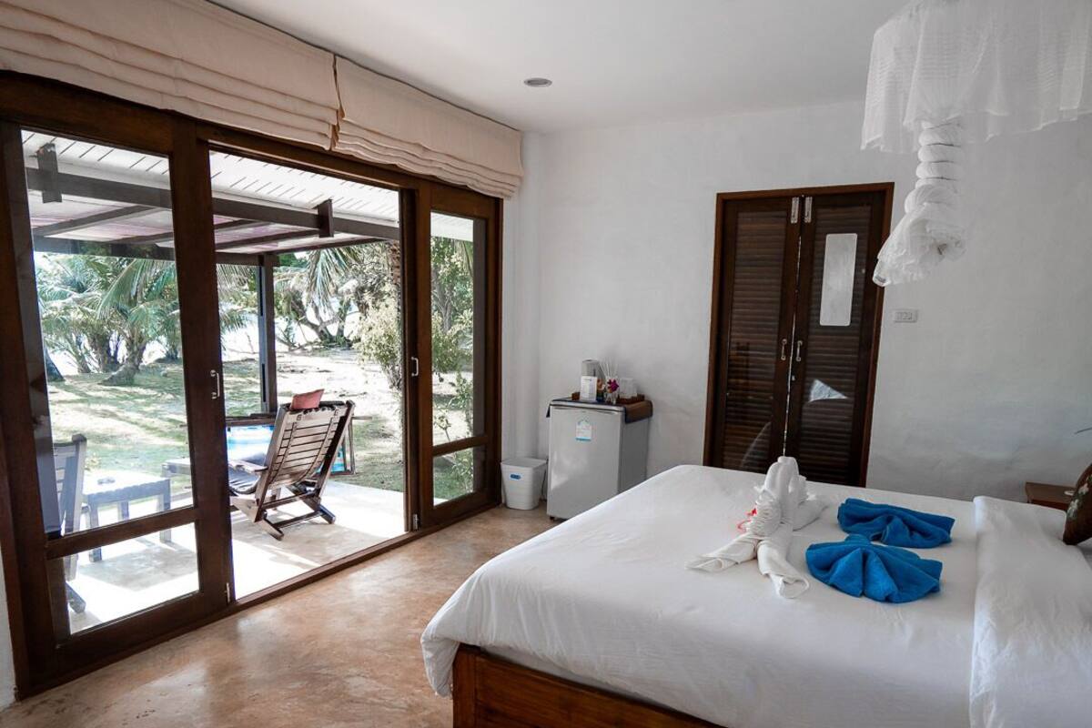 A bedroom with a large bed, a wardrobe, a small fridge, and a view of a garden through a sliding door.