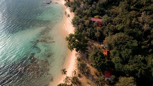 An aerial view of a beach with clear turquoise waters, a sandy shore, and a dense cluster of red-roofed buildings surrounded by lush greenery. - Lazy Day The Resort (Ko Mak)