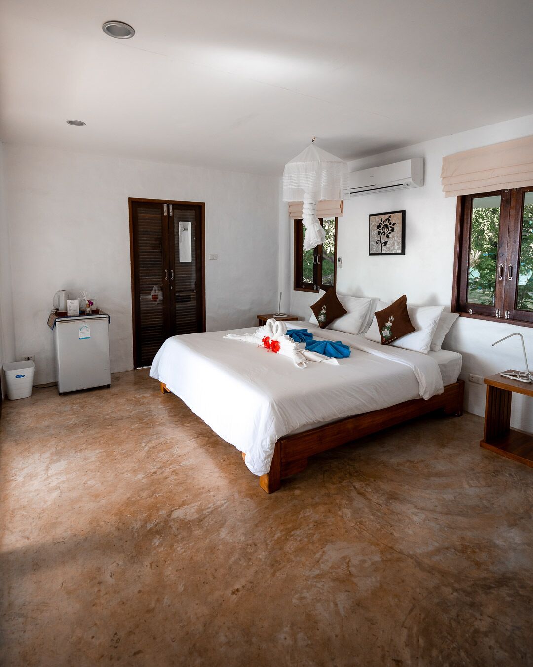 A bedroom with a large bed, a small bedside table, a wardrobe, a ceiling fan, and a window with a view of trees.