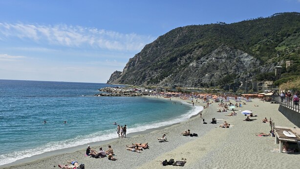 A beach with people sunbathing and swimming, a rocky hillside, and a clear sky. - Monterosso Apartment (Monterosso al Mare)