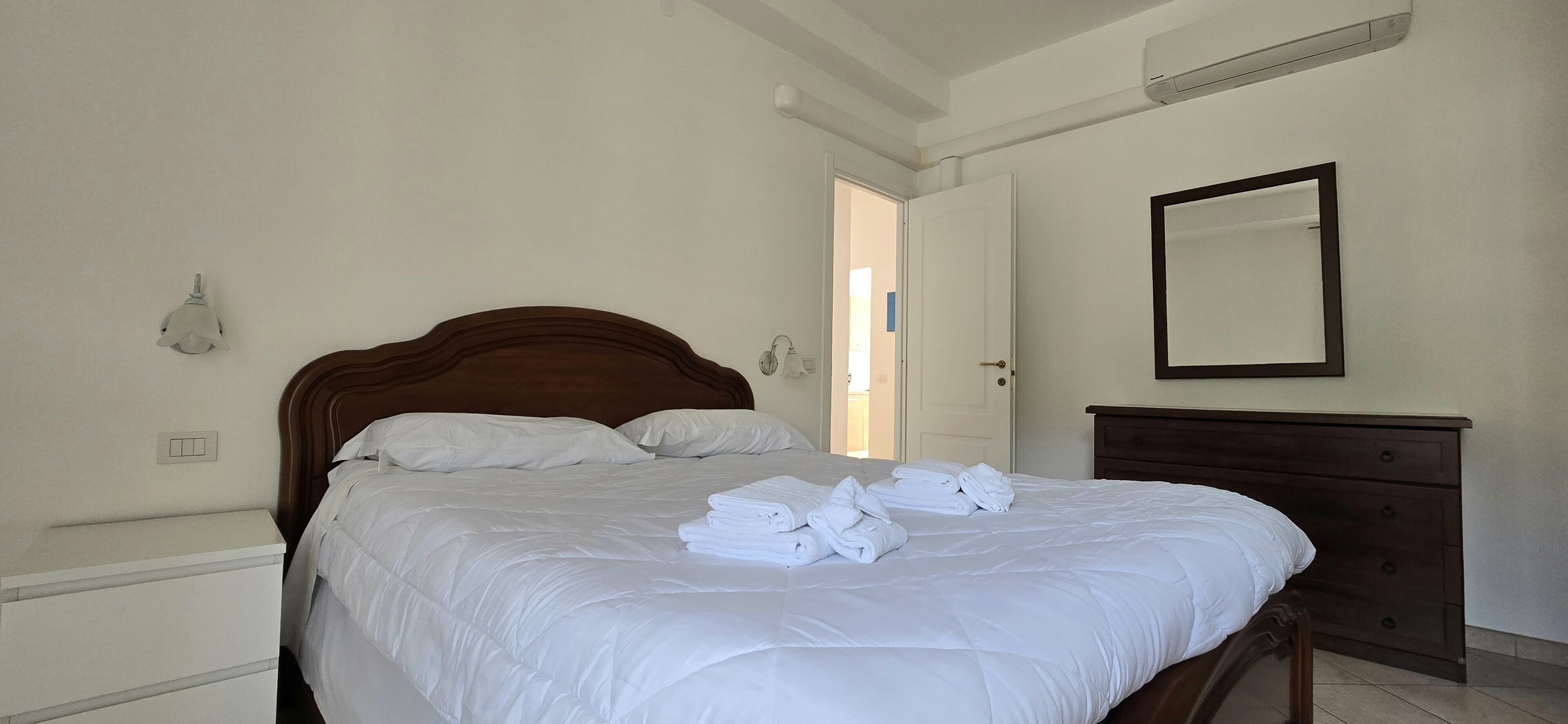 Una cama bien hecha con ropa blanca, un cabecero de madera, un aparador con cajones, un espejo fijado a la pared y un interruptor de luz en la pared.