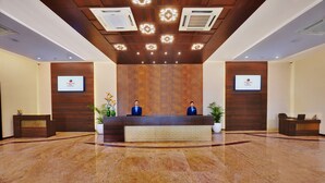 A reception area with a wooden reception desk, two receptionists, and two screens displaying the hotel’s logo. - Manpho Bell Hotel & Convention Center (Bengaluru)