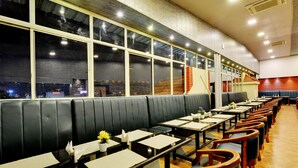 A restaurant with long black booths and wooden tables, large windows, and a city view. - Manpho Bell Hotel & Convention Center (Bengaluru)