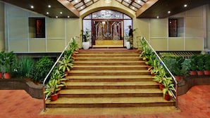 A grand staircase with a wooden handrail, flanked by potted plants, leading to a room with a large window and a decorative ceiling. - Manpho Bell Hotel & Convention Center (Bengaluru)