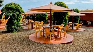 An outdoor seating area with round tables and chairs, covered by umbrellas, surrounded by gravel and greenery.