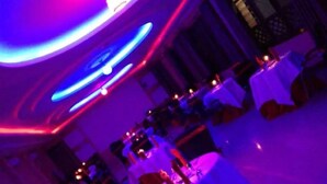 A dimly lit banquet hall with round tables covered in white tablecloths and red centerpieces.