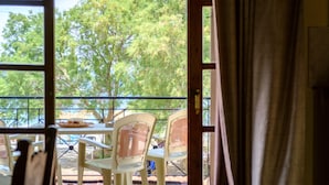 An outdoor seating area with a table and chairs, a view of trees through a window, and a curtain partially covering the window. - Seafront Paradise with sand Yard 10 m. from Beach! (Platanias)