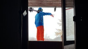 A person in a blue jacket and red pants standing on a wooden deck, holding skis. - La Grande Ourse (Valtournenche)
