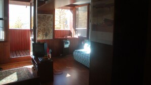 A cozy cabin interior with a wooden floor, a bed with a blue patterned bedspread, a desk with a computer, and a large window with a view of trees. - La Grande Ourse (Valtournenche)