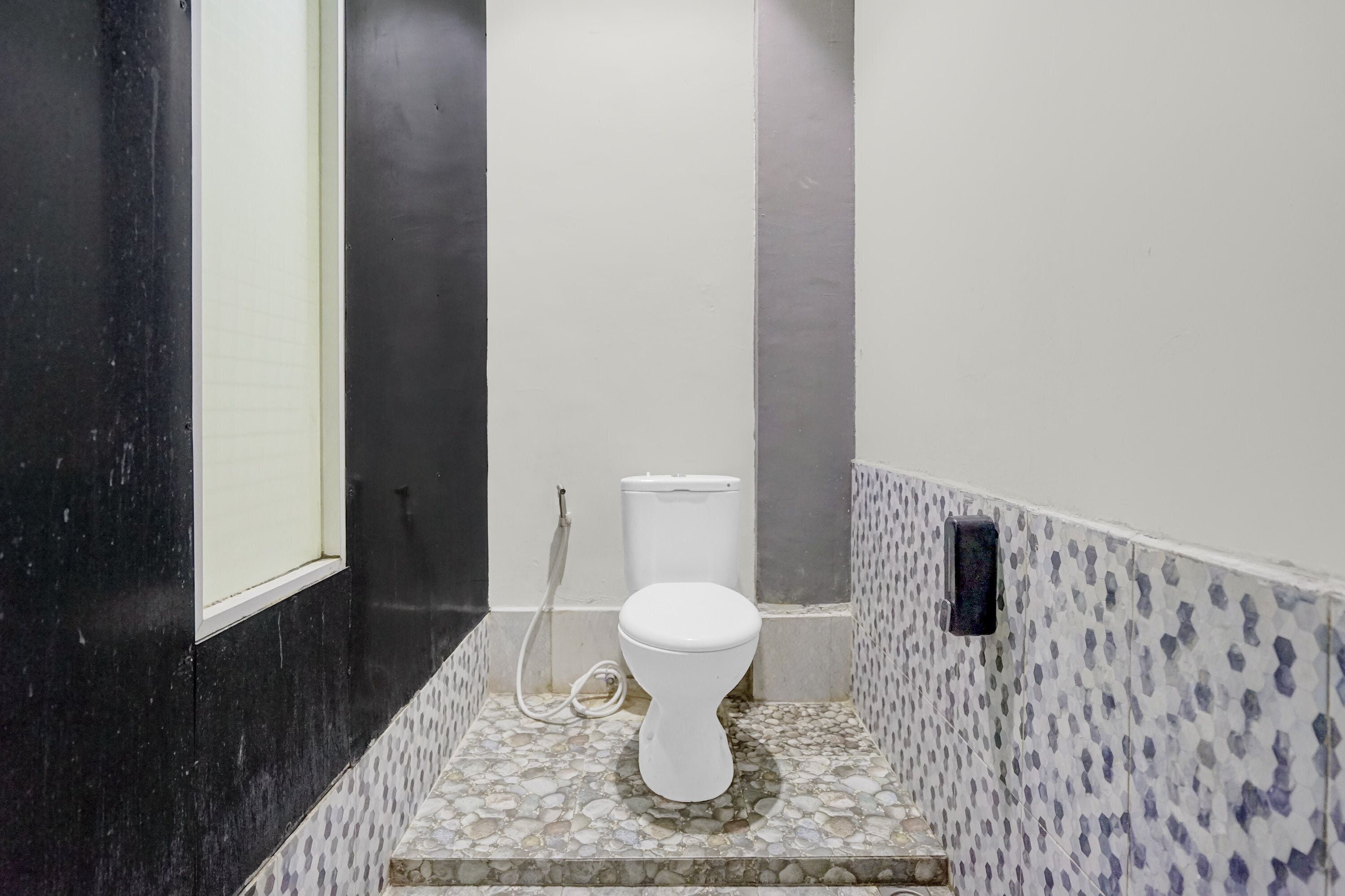 Une salle de bain avec des toilettes blanches, un sol carrelé, un mur noir et un mur blanc avec une petite fenêtre.