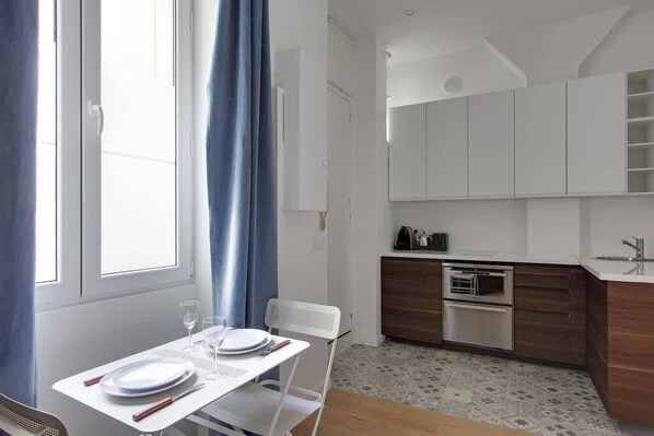 A modern kitchen with white cabinets, a built-in oven, and a sink. There is a dining area with a table set for two, and a window with blue curtains.