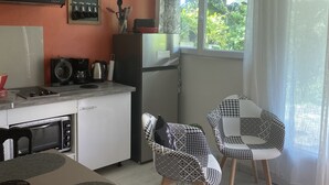 A compact kitchen with a microwave, stove, and refrigerator. There are two patterned chairs and a dining table. A window with curtains provides natural light. - Sweet Pea F2 flirtatious in the countryside (Fort de France)