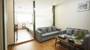 Un salon moderne avec un canapé gris, une table basse et une plante en pot. On aperçoit un lit au fond et une porte qui donne sur une autre pièce.