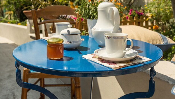Ein blauer runder Tisch mit Teekanne, Tasse und Glas, draußen aufgestellt, mit Stuhl und Pflanzen im Hintergrund.