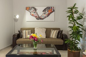 A living room with a sofa, a glass coffee table with a vase of flowers, a painting on the wall, and a potted plant.