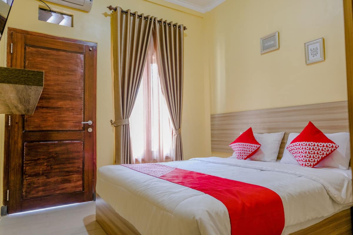 A hotel room with a wooden door, a bed with white bedding and red accents, and two framed pictures on the wall.