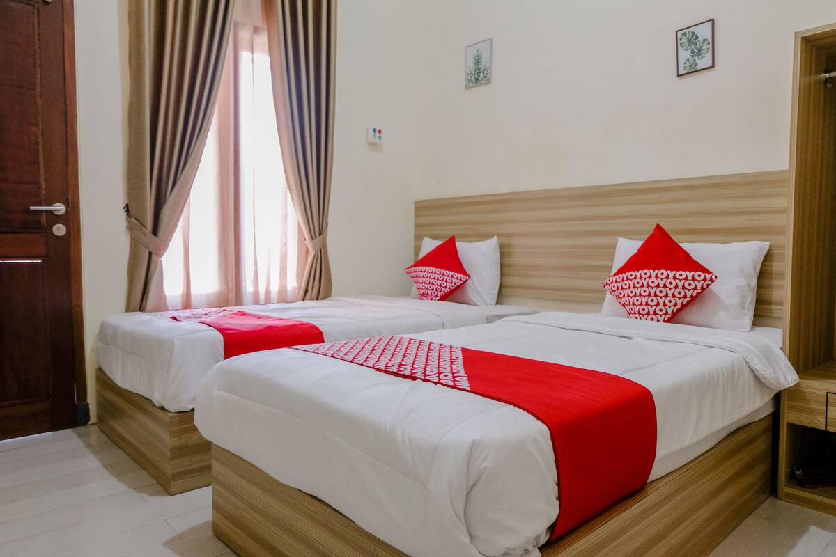 A hotel room with two beds, each with white bedding and red accents, a wooden headboard, and a window with curtains.