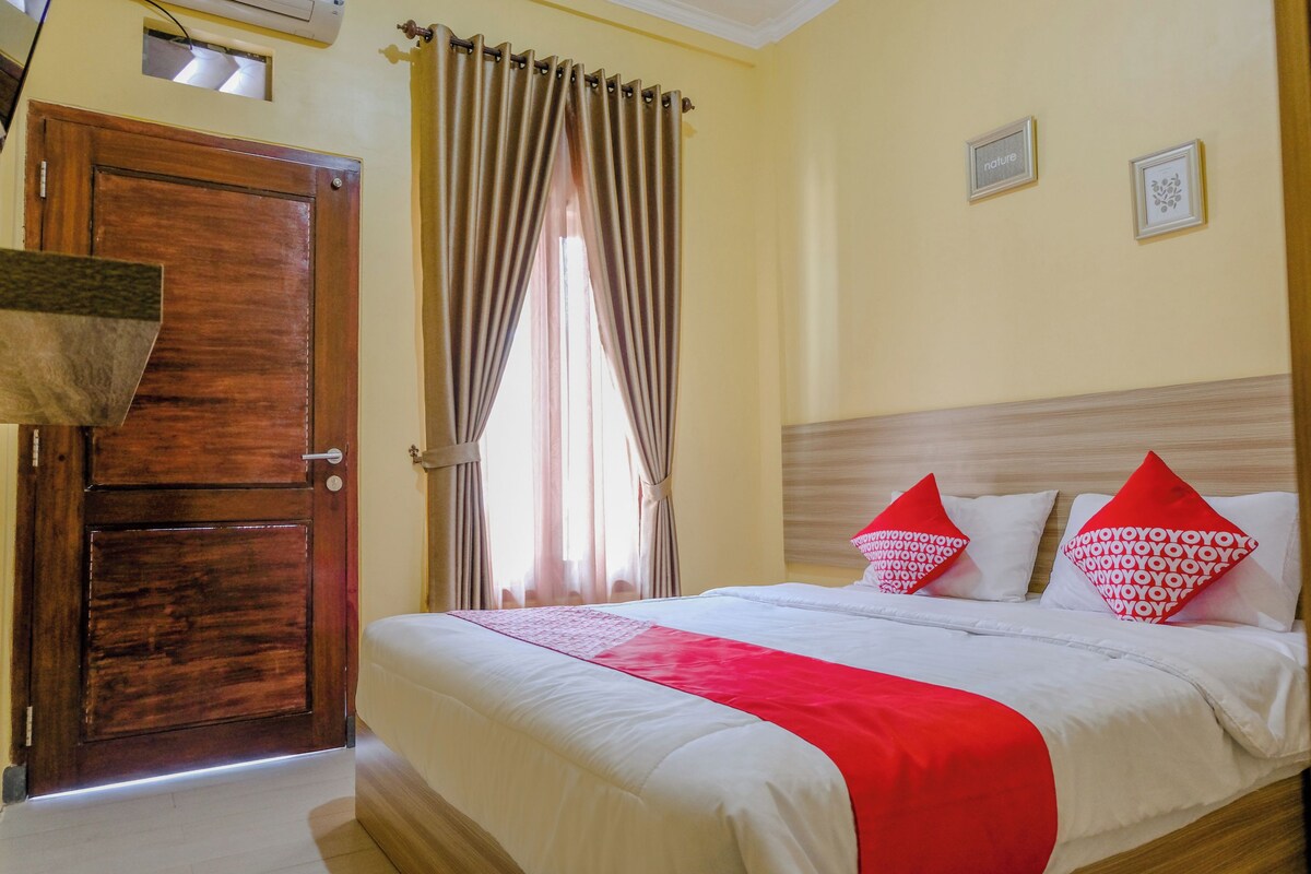 Ein Hotelzimmer mit einer Holztür, einem Bett mit weißer Bettwäsche und roten Kissen, einem Fenster mit Vorhängen und einer Wand mit zwei gerahmten Bildern.