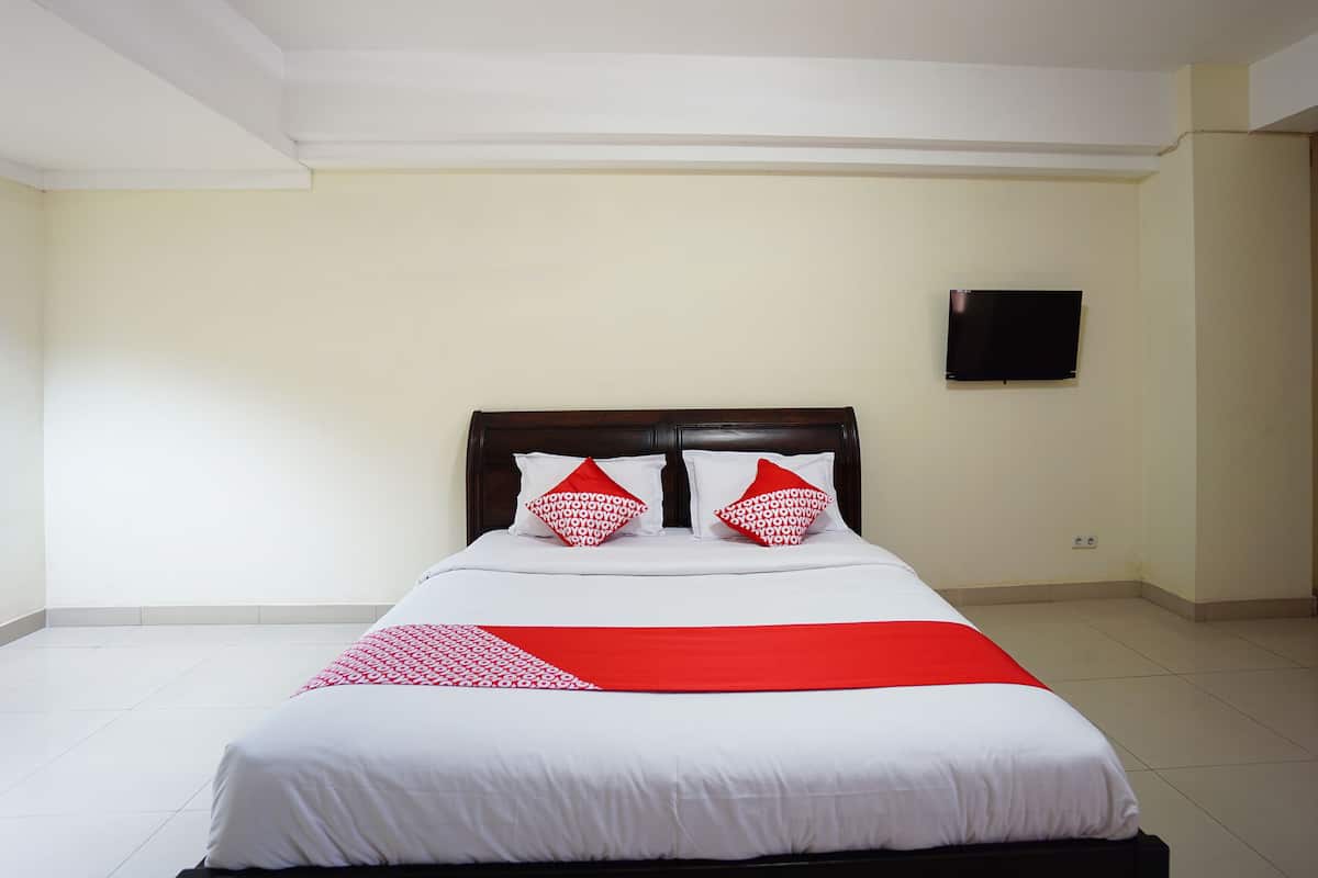 A neatly made bed with white and red bedding, a wooden headboard, and a flat-screen TV mounted on the wall.