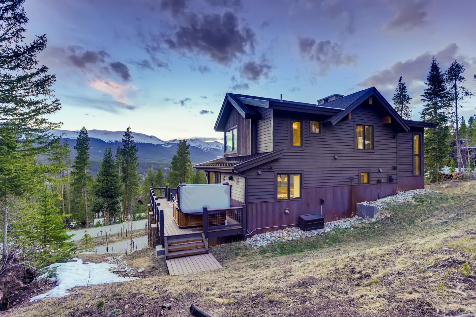 A two-story wooden house with a deck, surrounded by mountains and trees.