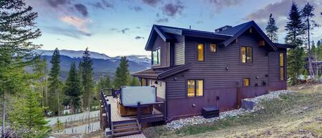 A two-story wooden house with a deck, surrounded by mountains and trees.