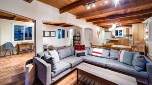 A modern living room with a grey sectional sofa, wooden flooring, and a kitchen area visible through an open archway. - 66SL - Hot Tub - WiFi - 3+ Bedroom - Sleeps 10 Home by RedAwning (Deming)