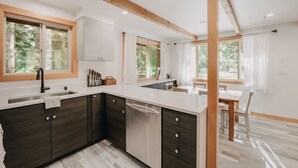 Une cuisine moderne avec des meubles en bois foncé, des plans de travail blancs et un lave-vaisselle encastré. De grandes fenêtres laissent entrer la lumière naturelle et offrent une vue sur l’extérieur.