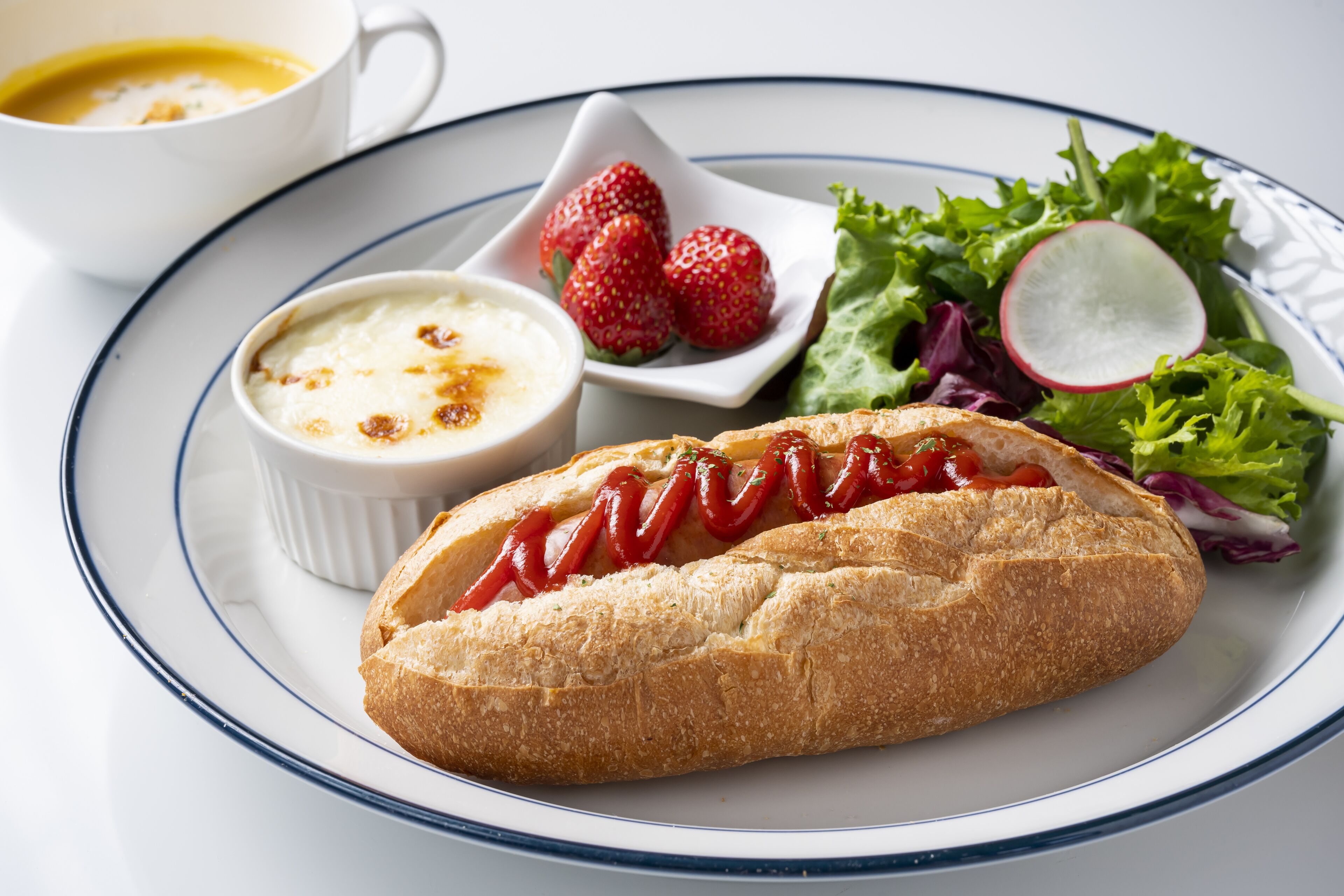A plate with a bread roll, a small bowl of soup, a cup of coffee, and a salad with strawberries and radish.