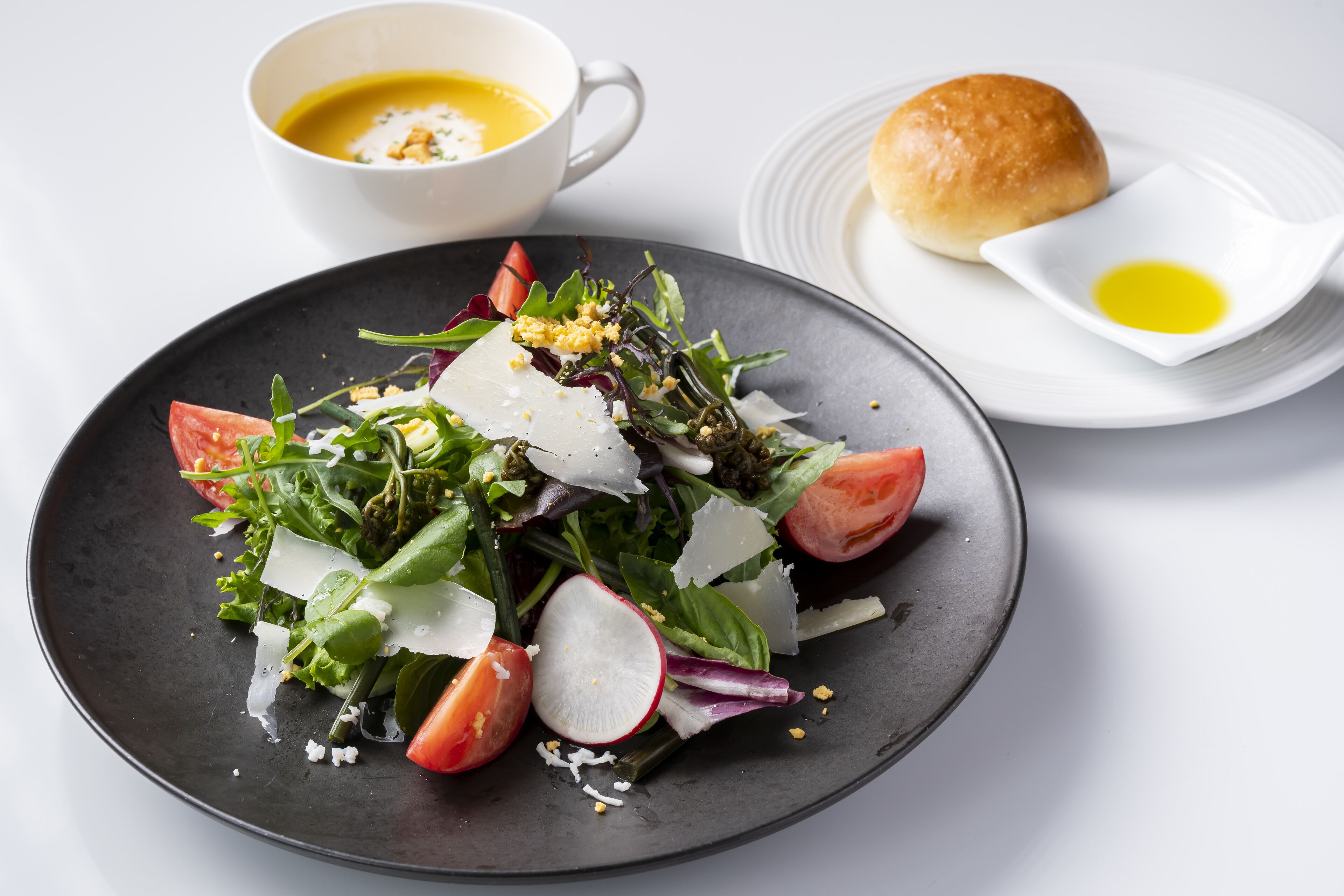 Un plato de ensalada con diversas verduras y vegetales, un tazón pequeño de sopa, un panecillo y una pequeña fuente con aceite.