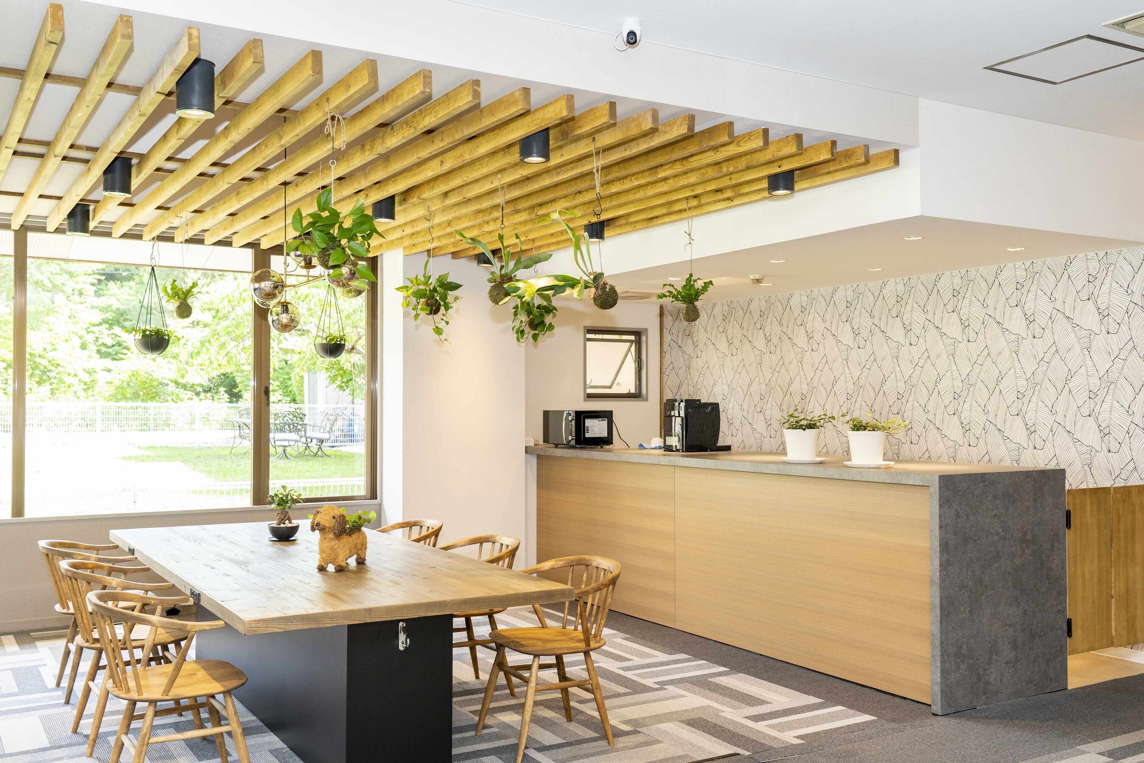 A modern kitchen with wooden beams, a wooden dining table with chairs, hanging plants, and a view of the outdoors.