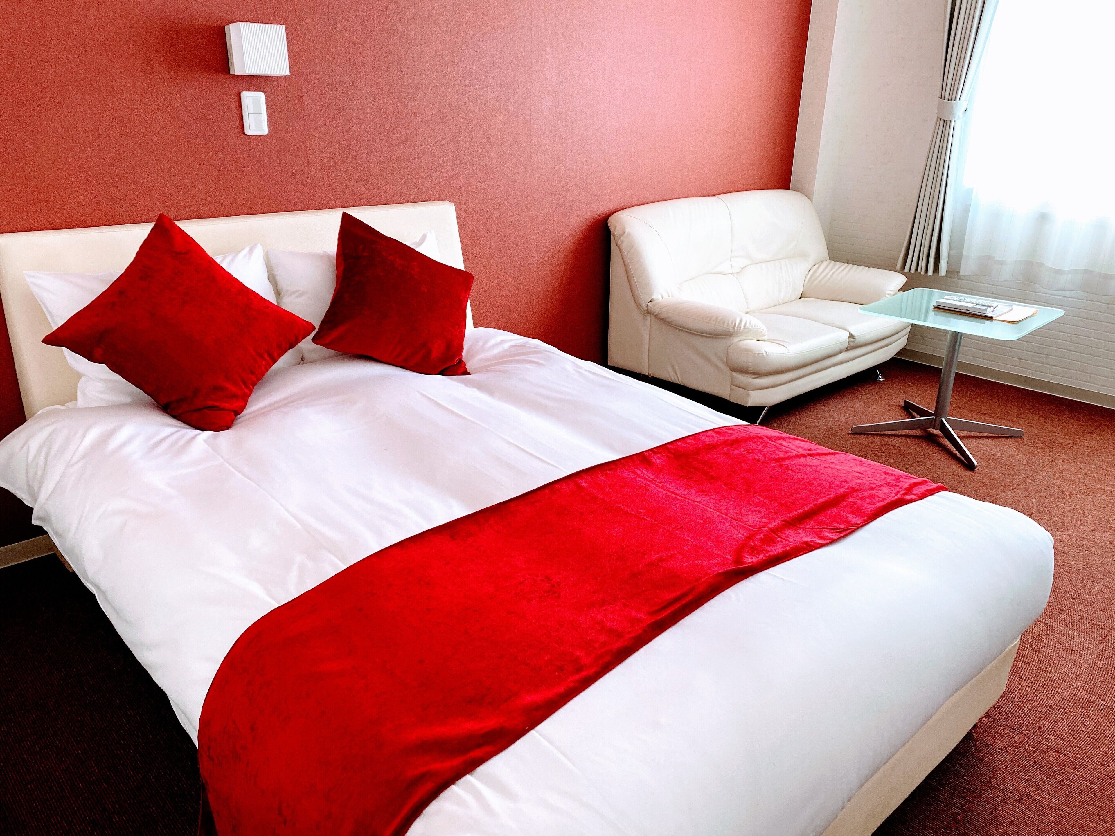 A hotel room with a bed, a white chair, a glass table, and a red accent wall.