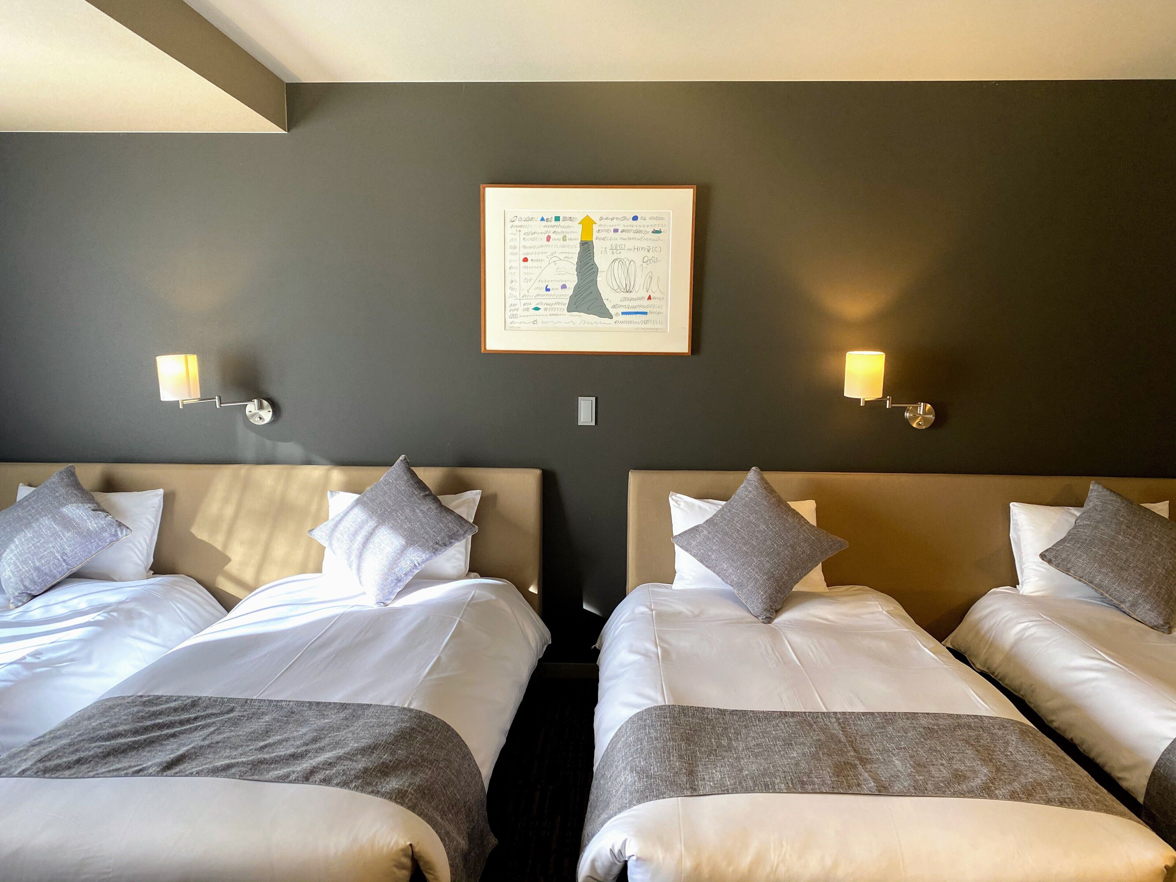 Habitación de hotel con dos camas, cada una con ropa blanca y almohadas grises. Hay lámparas de pared a cada lado de una obra de arte enmarcada sobre las camas.