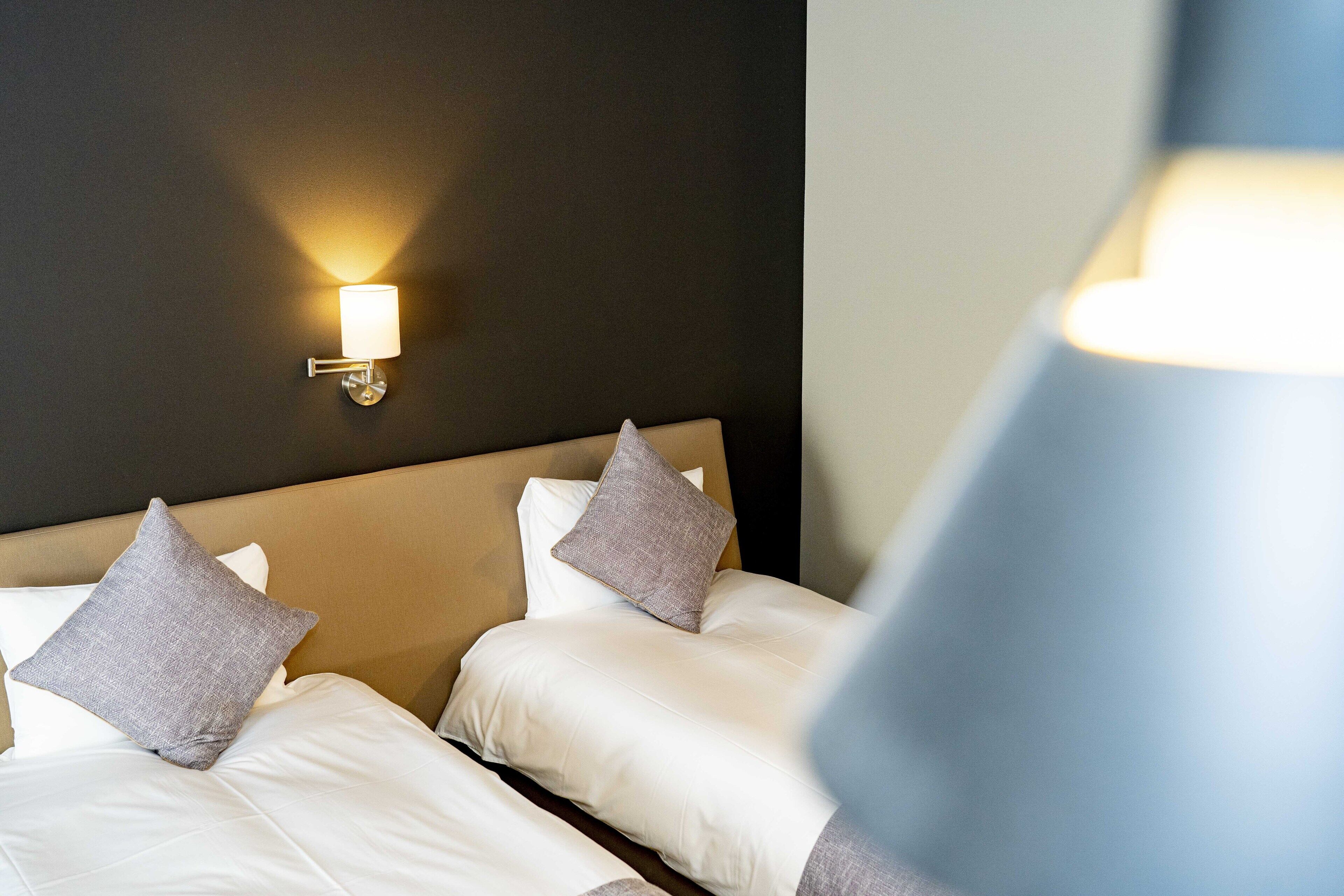 Habitación de hotel con dos camas, cada una con ropa blanca y almohadas grises, y una lámpara fijada a la pared.