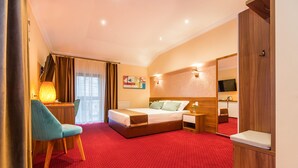 Una habitación de hotel con alfombra roja, una cama con ropa blanca, una mesita de noche de madera, una lámpara, una silla, un televisor y un espejo.