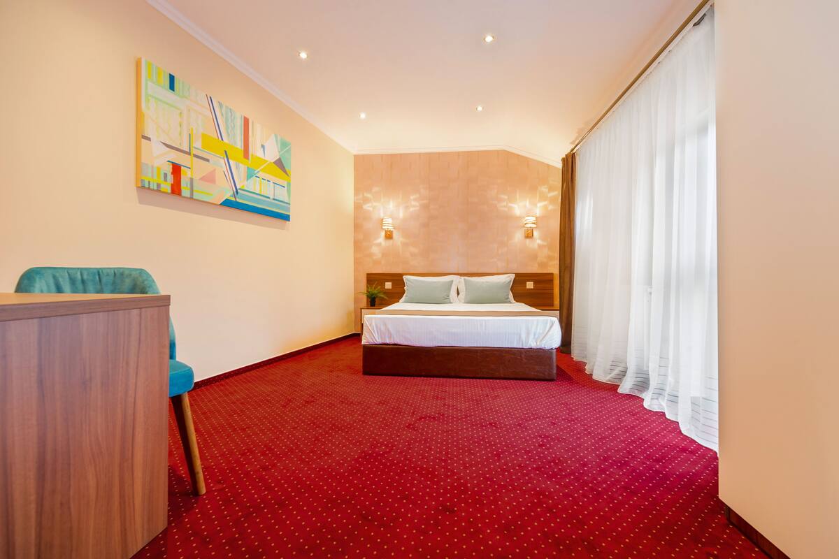 A hotel room with a large bed, a desk, a chair, a painting on the wall, and a window with curtains.