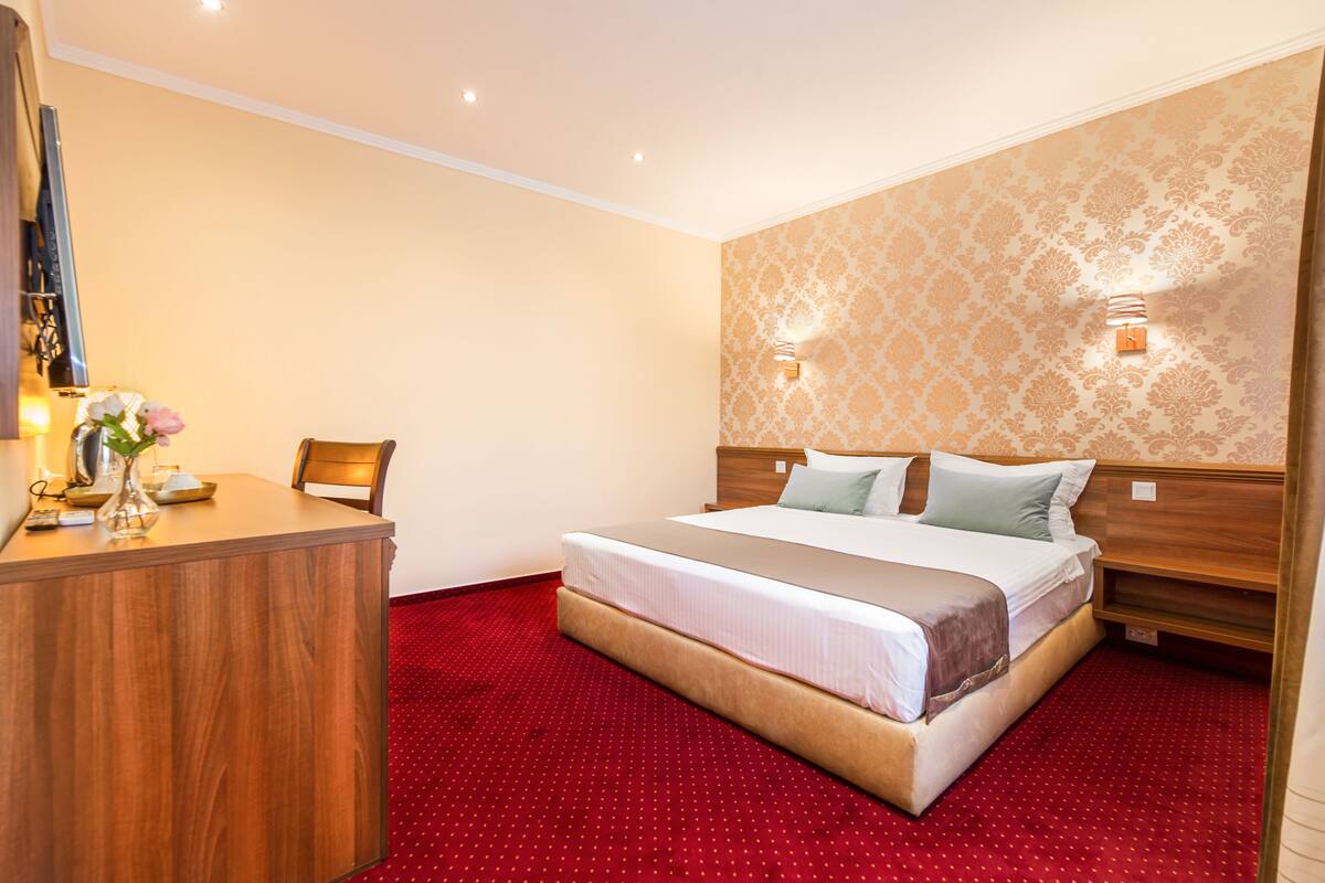A hotel room with a large bed, a wooden desk, and a red carpeted floor.