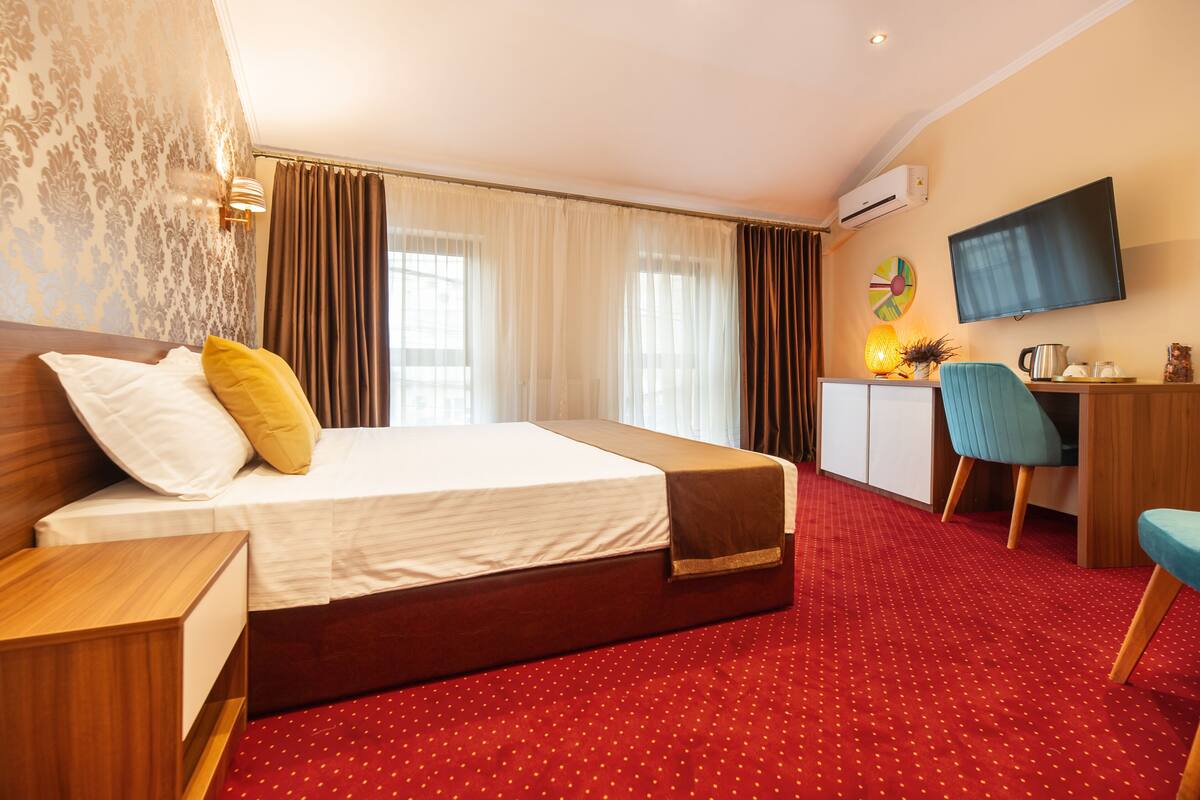 A hotel room with a bed, a desk, a chair, a TV, and a red carpet.