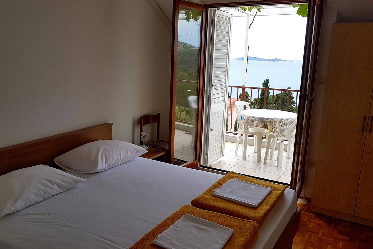 A bedroom with a bed, a nightstand, a chair, and a view of an outdoor balcony with a table and chairs.