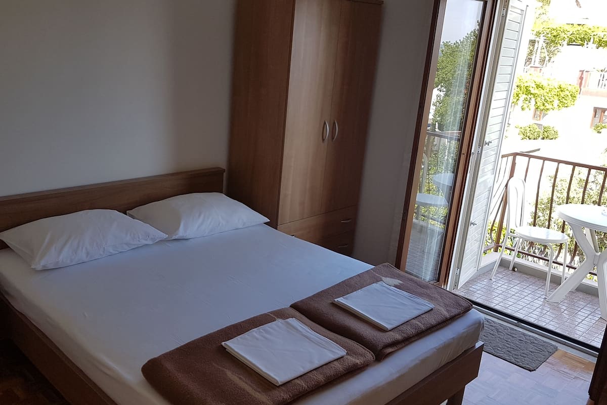 A bedroom with a bed, a nightstand, a wardrobe, and a balcony with a table and chairs.