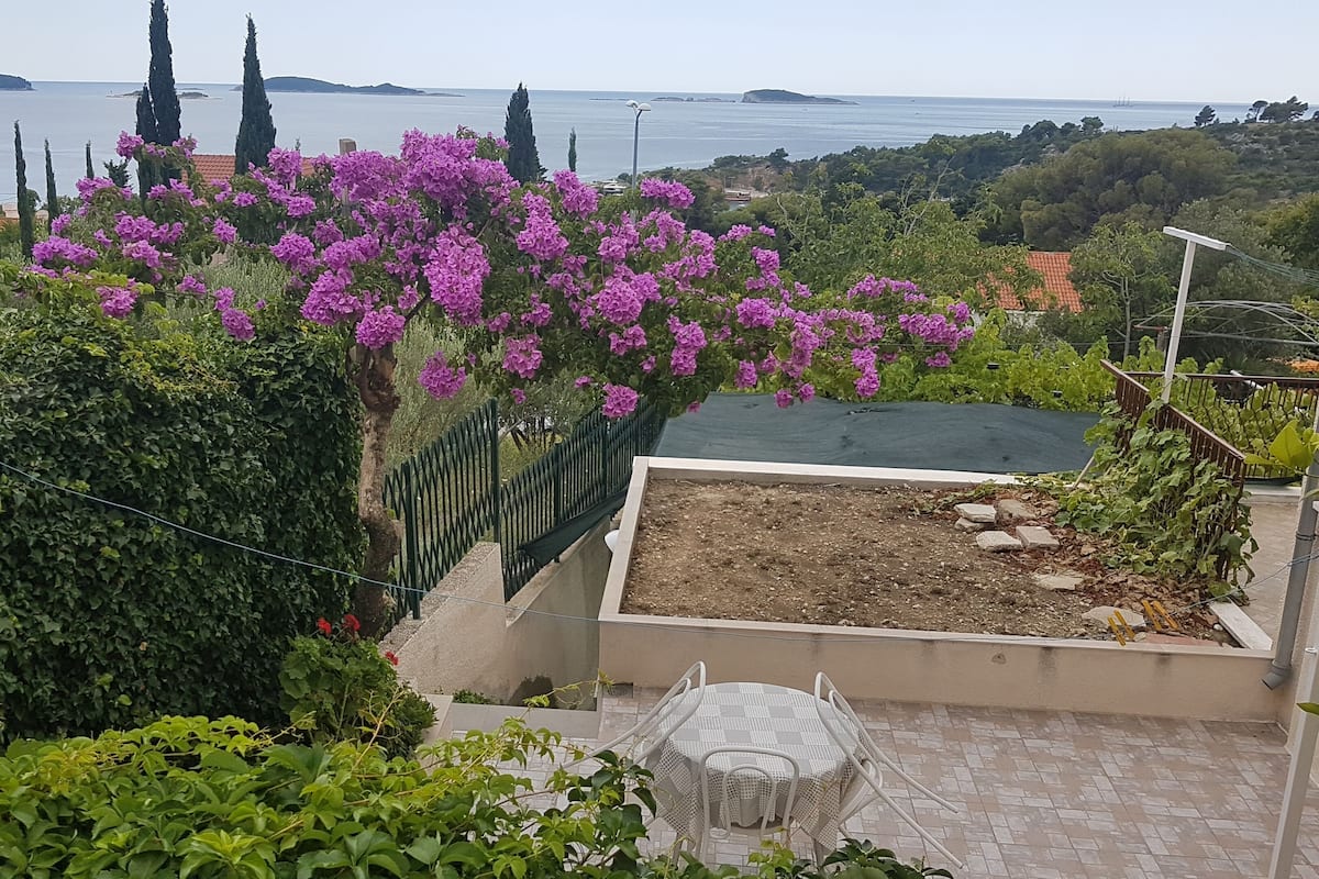 A garden with a pergola covered in purple flowers, a paved patio with a white chair, and a view of the sea and distant hills.