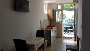 A compact kitchen with a dining area, a TV mounted on the wall, and a view of the outside through a door. - Apartments Kust (Zupa dubrovacka)